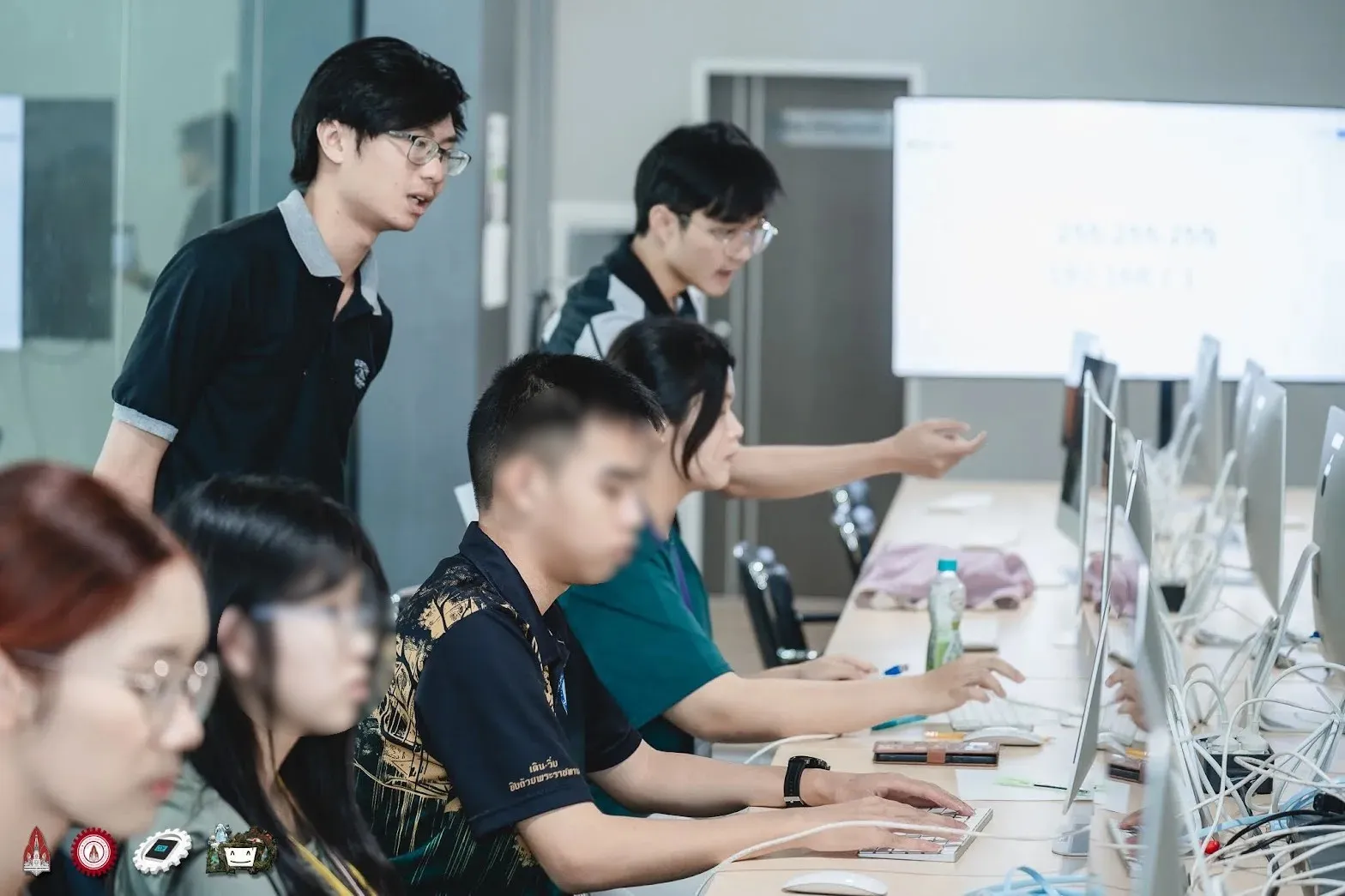 Staff pointing at a row of iMacs with network cables while campers type; an IP address and subnet mask on the screen behind